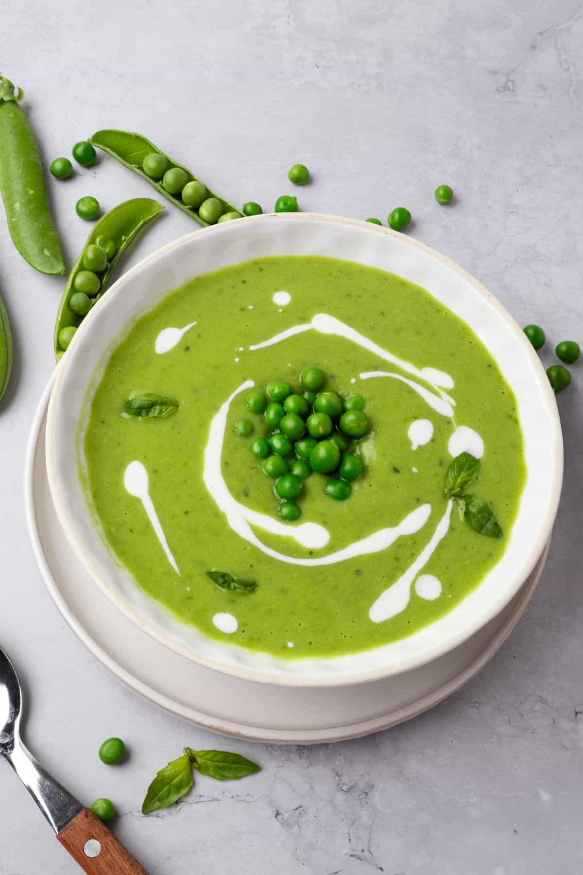 A bowl of green pea soup on the table drizzled with yogurt and topped with blanched peas and fresh basil leaves.