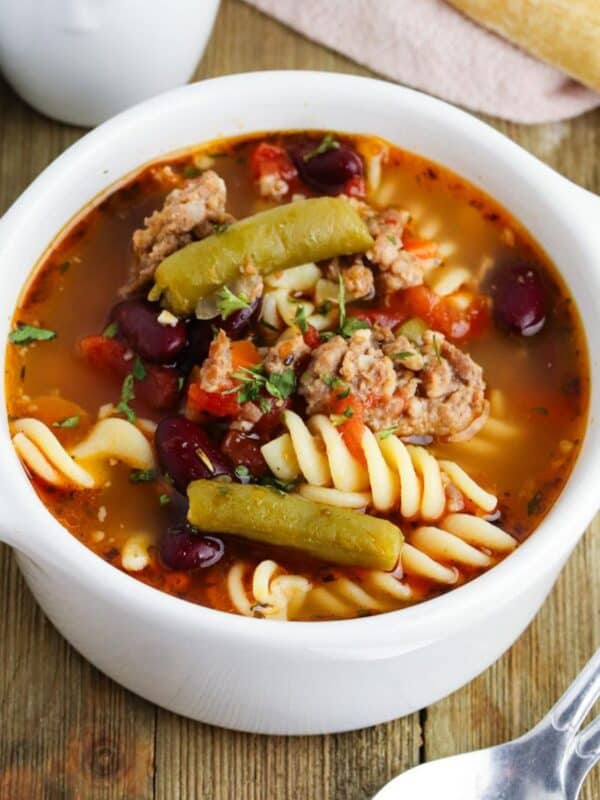 A small white crock of Italian sausage soup with pasta on a wooden table with spoons next to it.