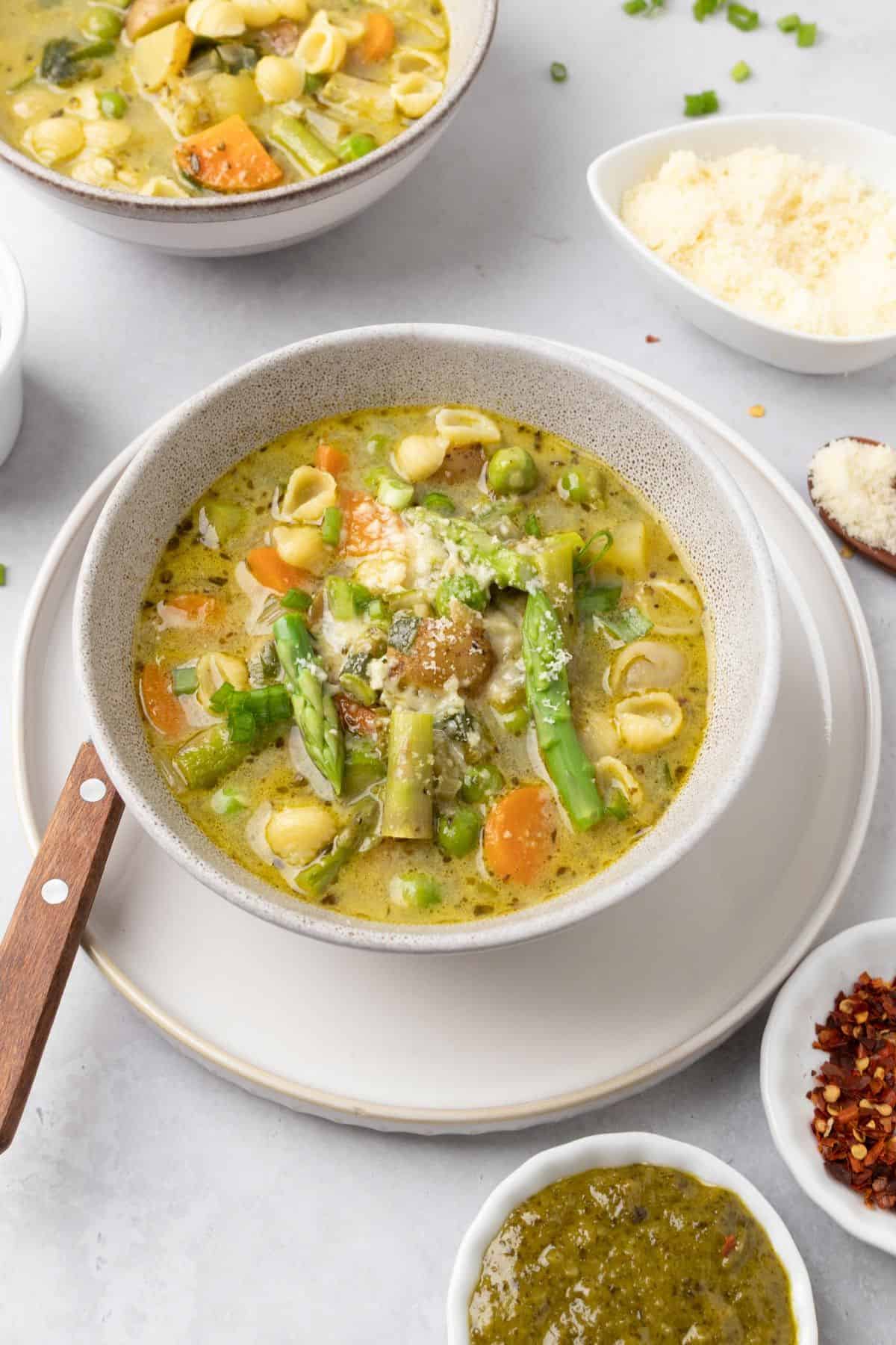 A bowl of green minestrone soup on the table with another bowl in the background.