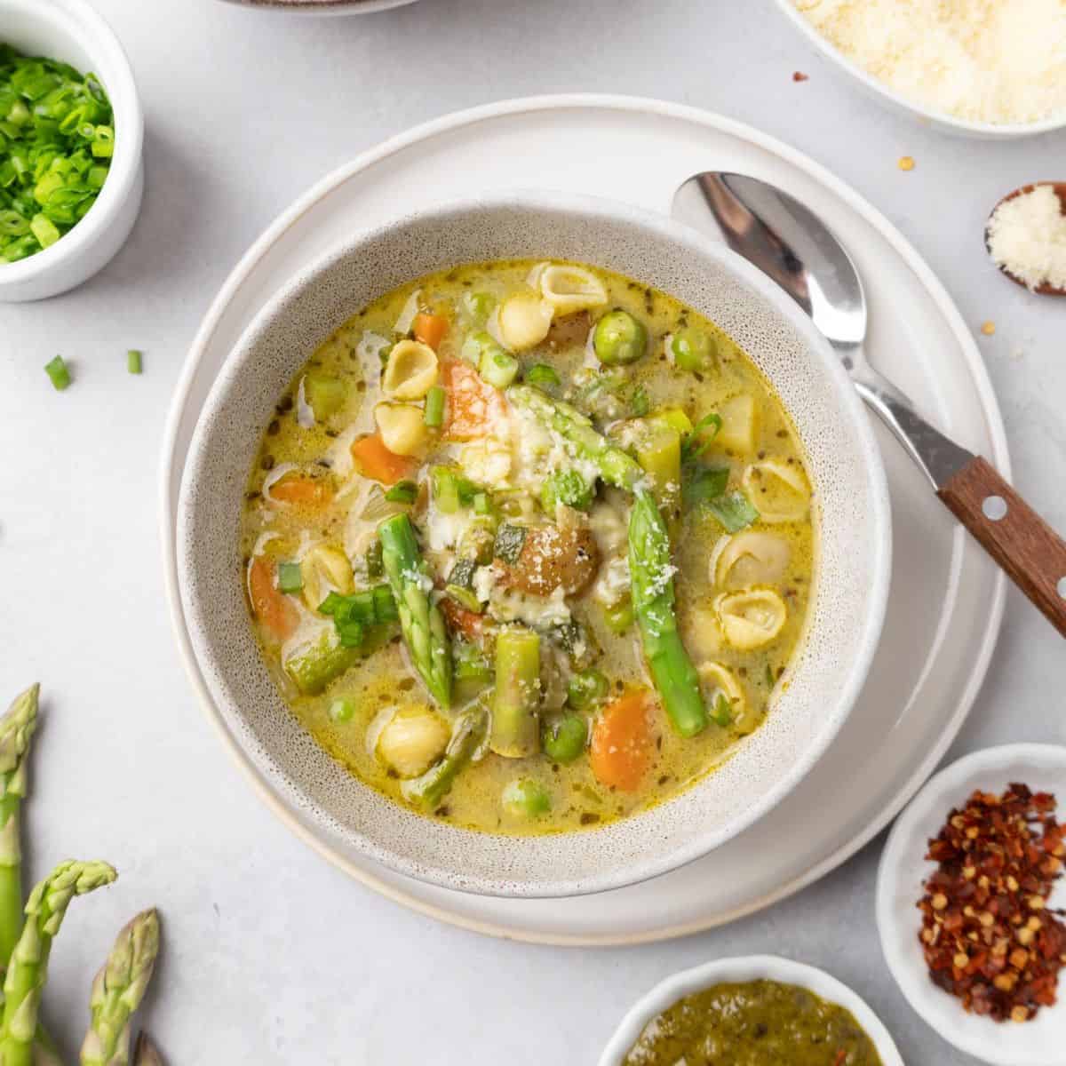 A bowl of spring minestrone soup on a plate with a spoon to the side.