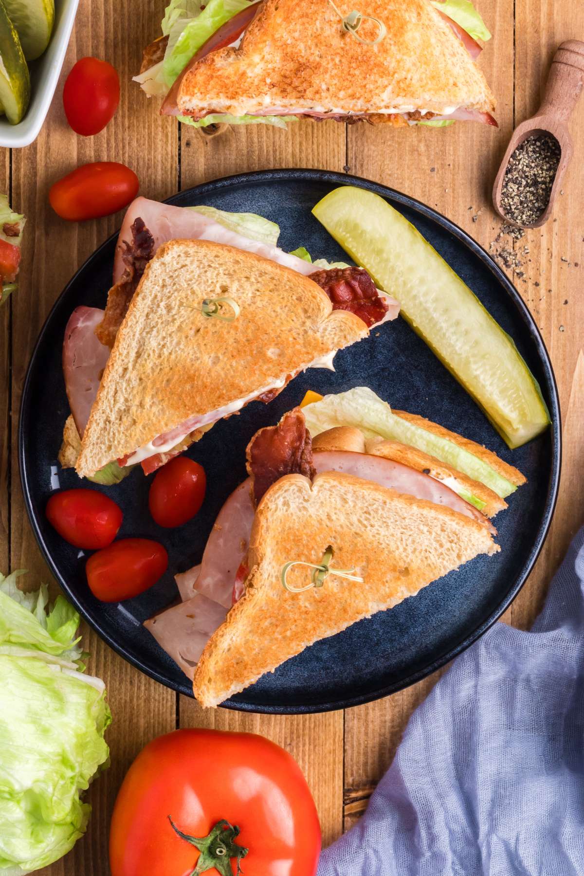 A chicken club sandwich on a plate with a pickle spear and grape tomatoes to the side.