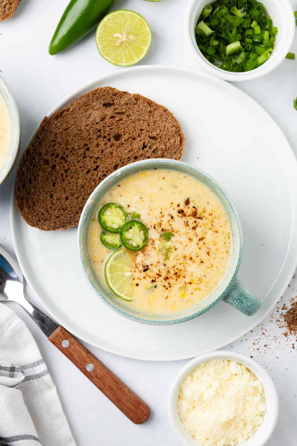 A bowl of Mexican street corn soup on a plate with a slice of dark bread to the side.
