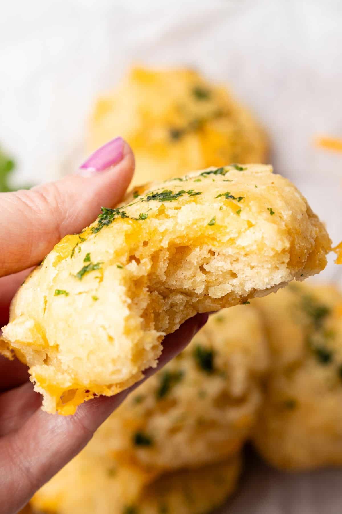 Bite taken from a cheese biscuit held up by a hand over the plate of more biscuits.