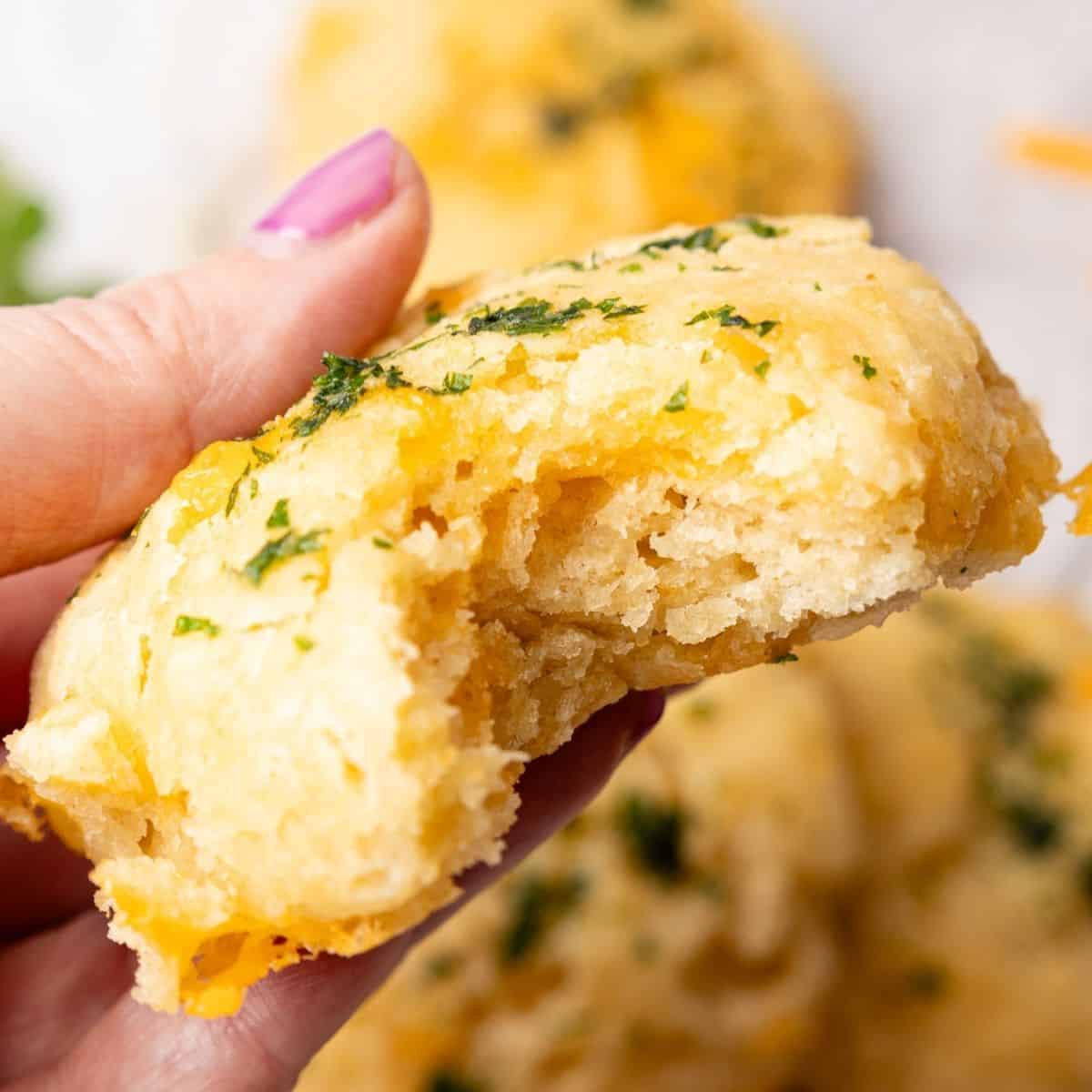 A hand holding up a cheddar biscuit with a bite missing.
