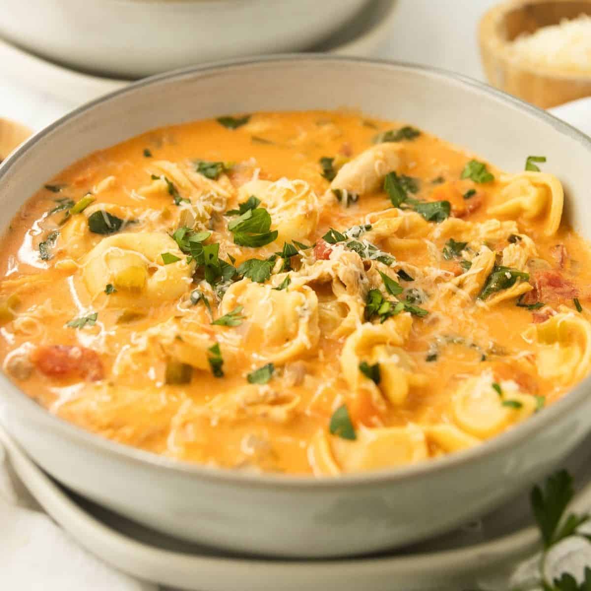 A bowl of chicken tortellini soup topped with chopped parsley and another bowl in the back.