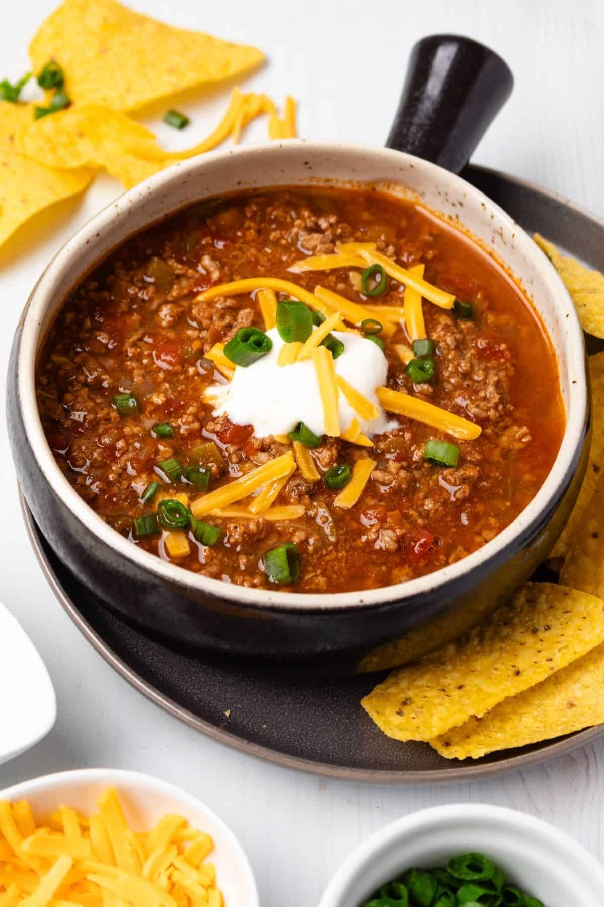 A bowl of beef chili without beans with chips, sour cream, cheese, and green onions.
