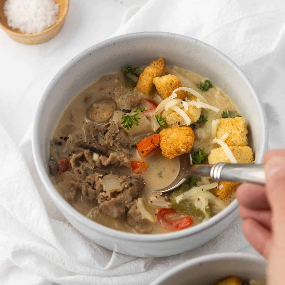 A bowl of Philly cheesesteak soup in a bowl with a hand holding a spoon in it.