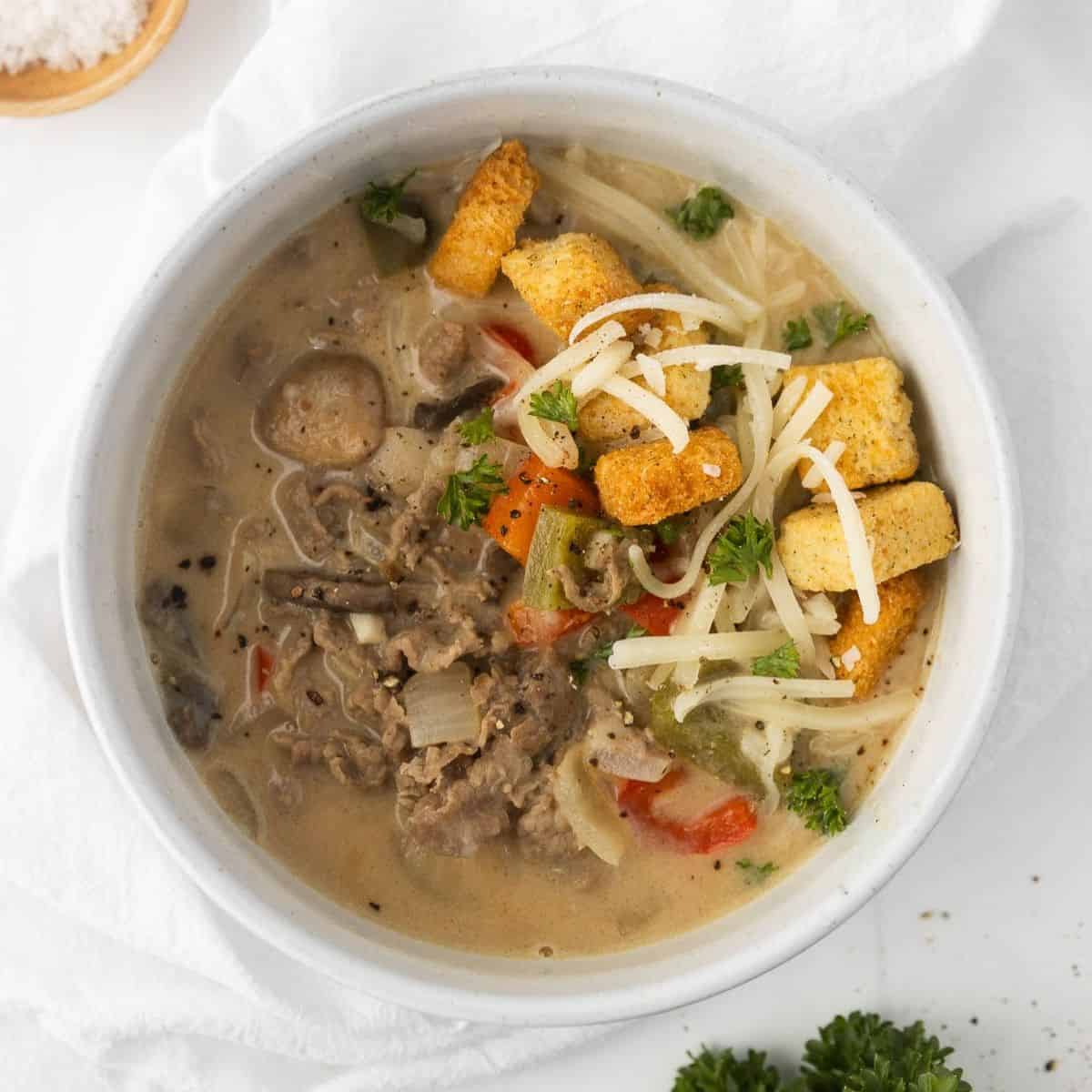 A bowl of Philly cheesesteak soup with shredded cheese and croutons on top.