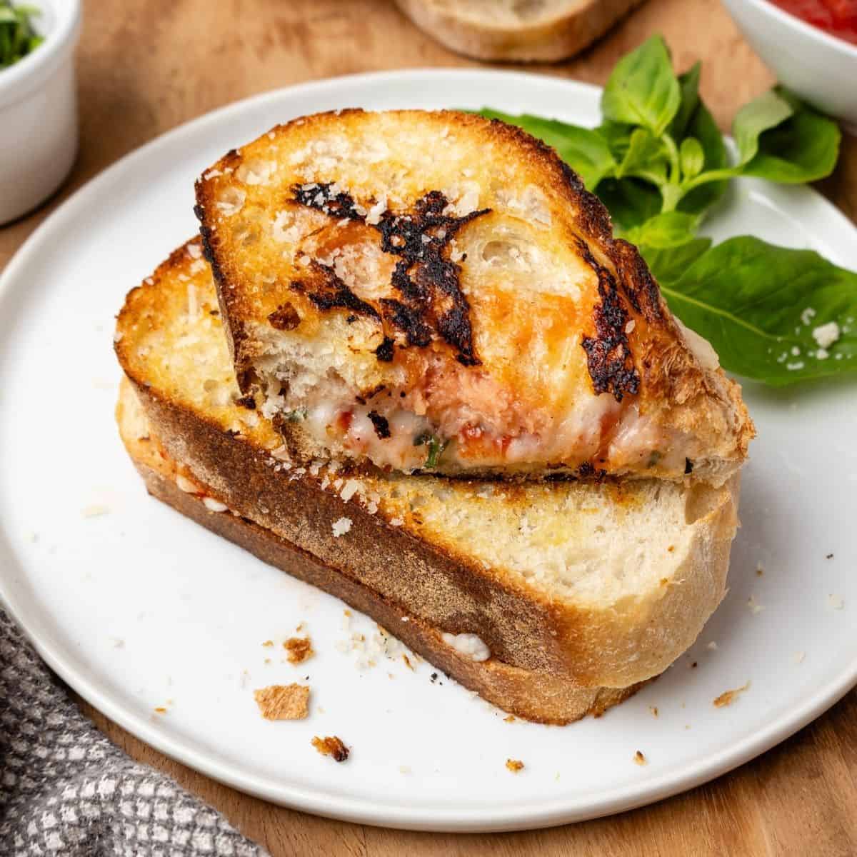 A pizza grilled cheese on a plate with a half of a sandwich on top and fresh basil in the background.