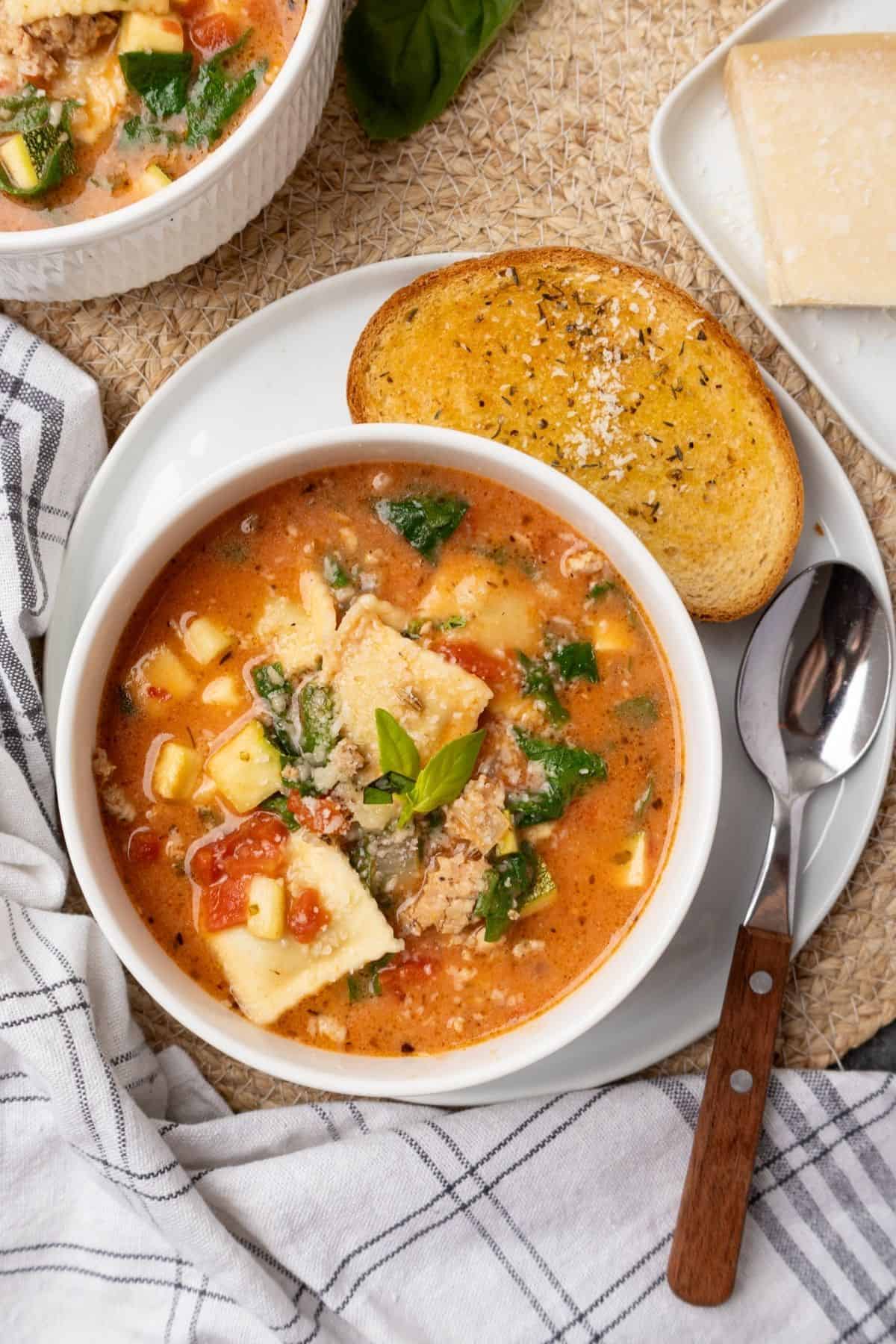 A bowl of ravioli soup on a plate with a piece of garlic bread. 