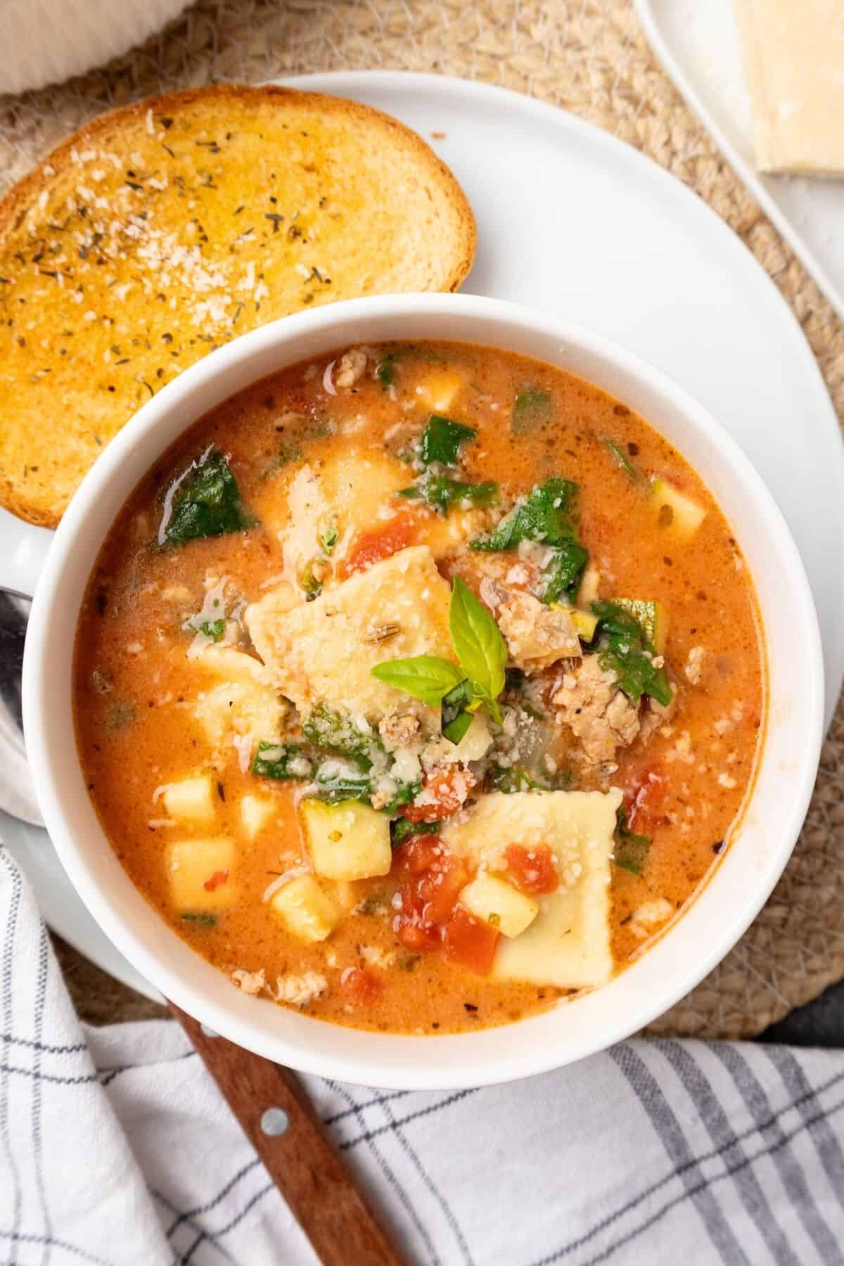 Ravioli soup in a white bowl on a plate with some garlic bread. 