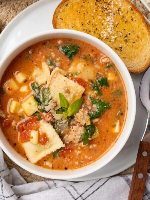A bowl of ravioli soup on a plate with garlic bread and a spoon to the side.