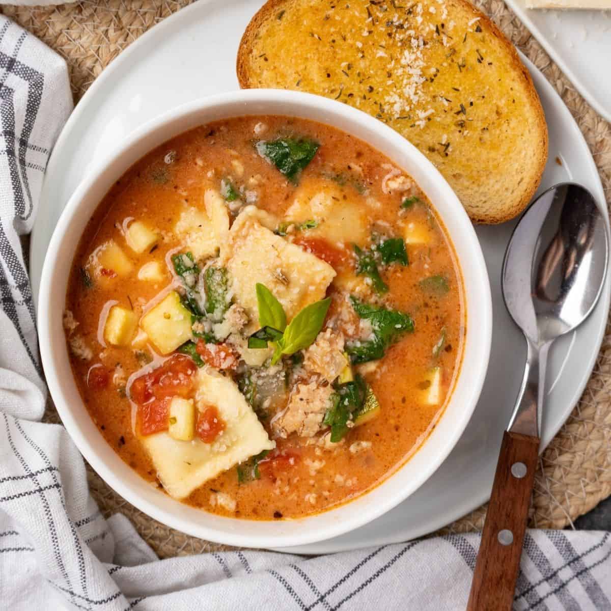 A bowl of ravioli soup on a plate with garlic bread and a spoon to the side.