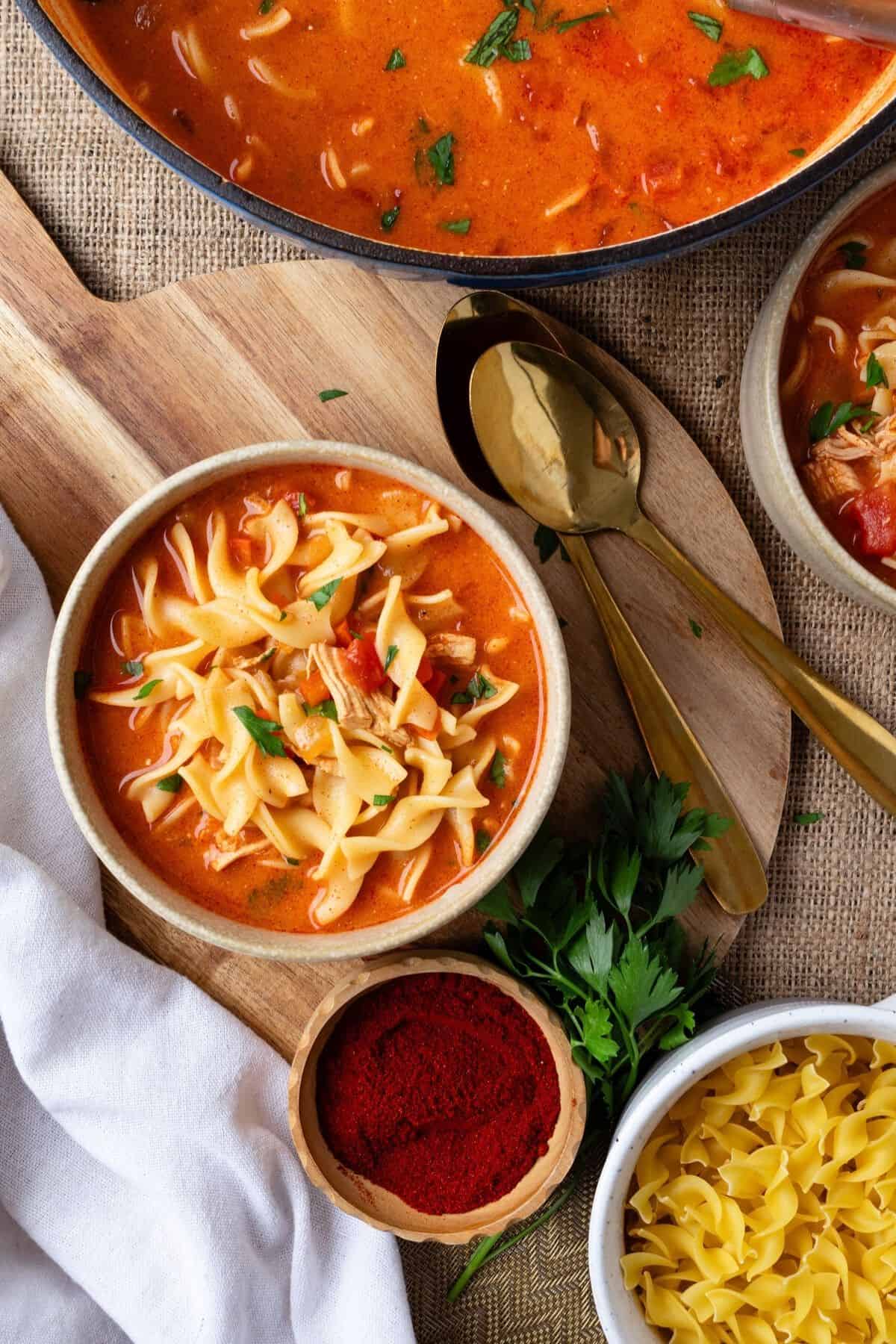 A bowl of chicken paprikash on a wooden board with a pot of more soup to the side.
