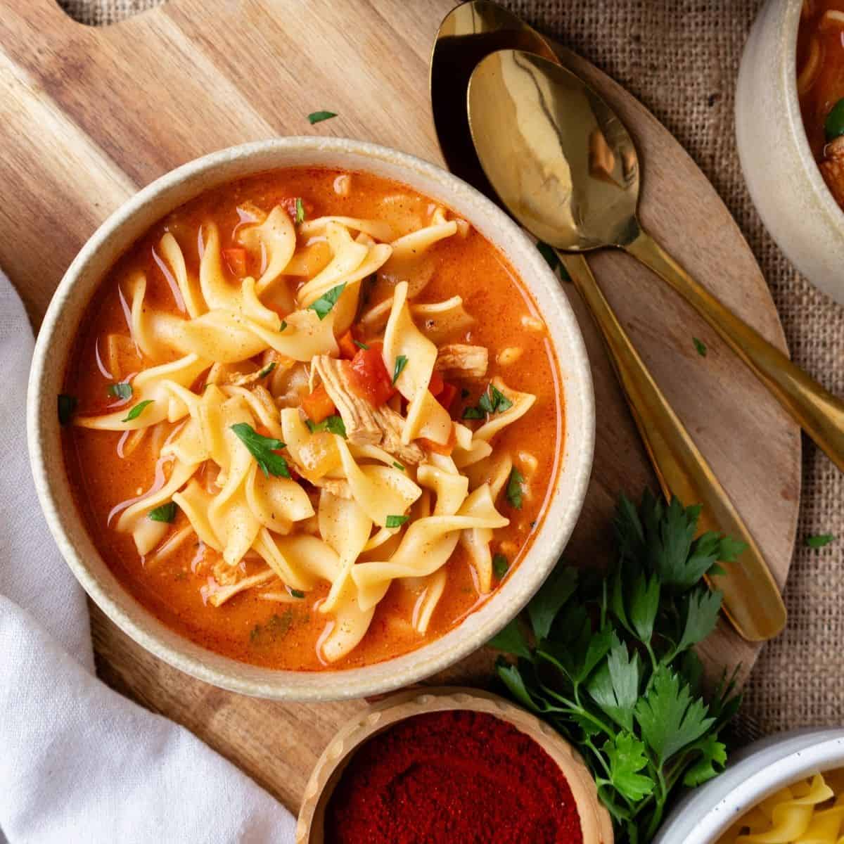 A bowl of chicken paprikash soup on a wooden surface with a spoon to the side.