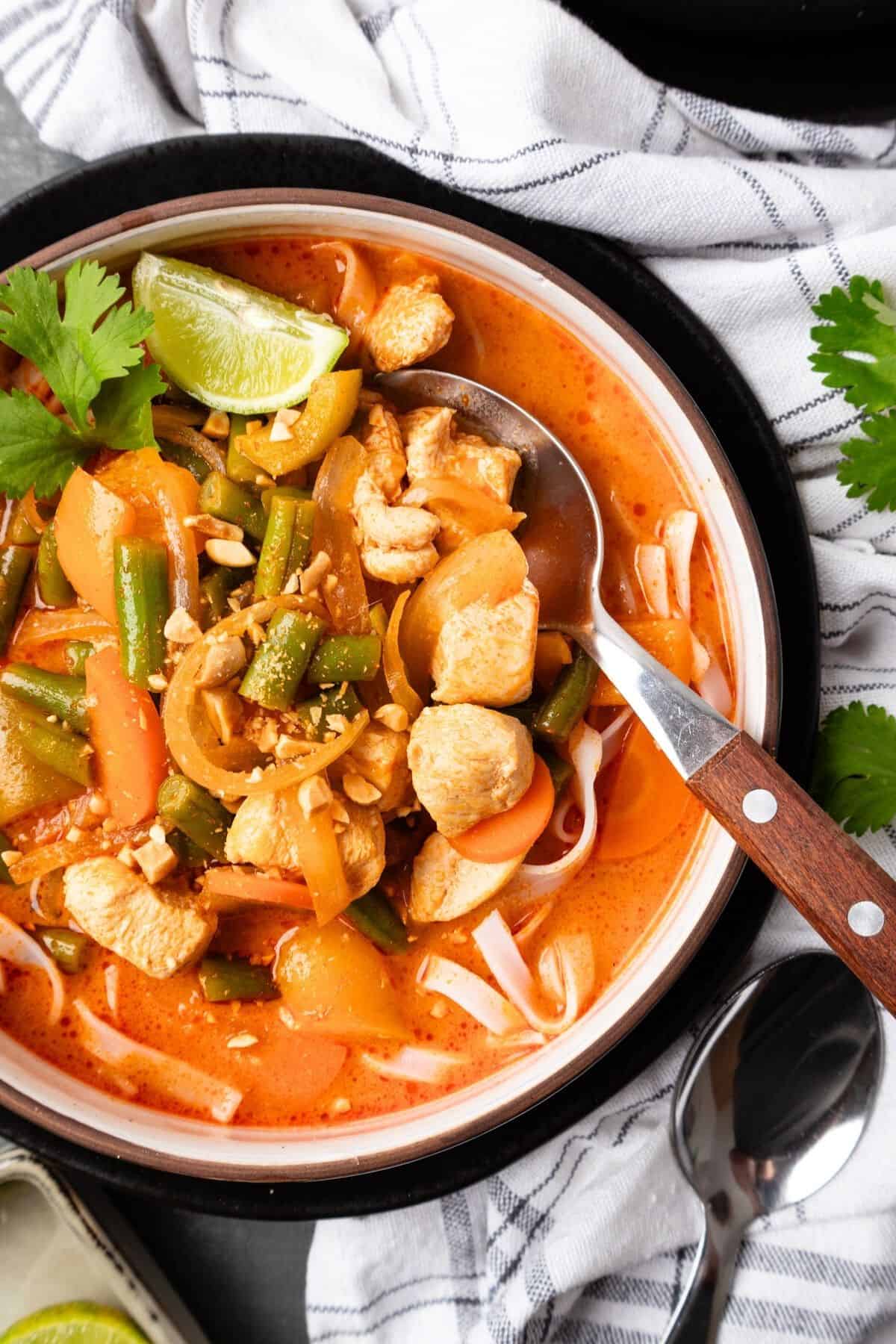 A bowl of Thai red curry soup with a spoon in the bowl and garnished with cilantro and lime.
