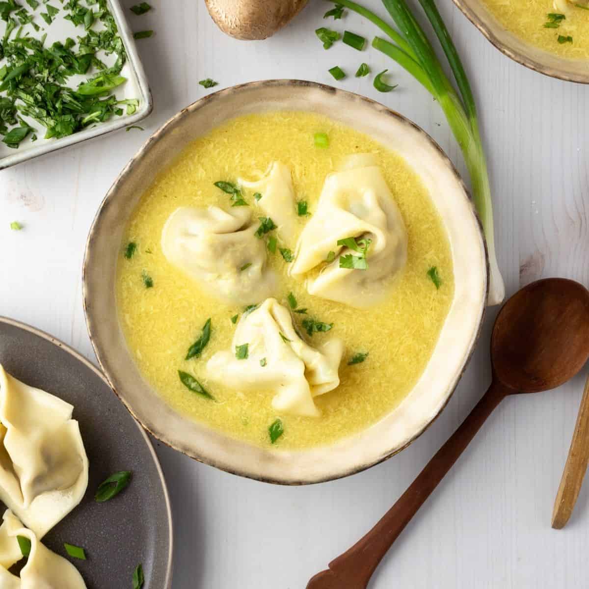 A bowl of wonton egg drop soup on a table garnished with green onions.