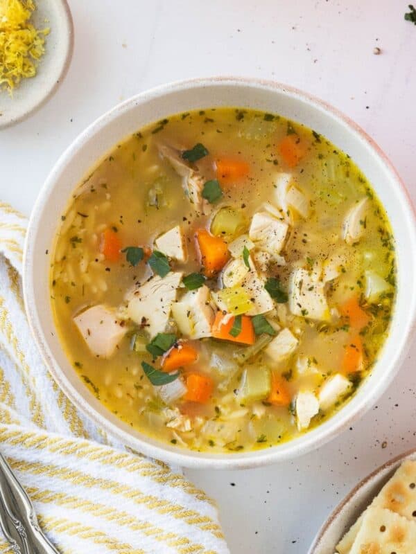 A bowl of lemon chicken orzo soup next to a yellow and white napkin with a spoon.