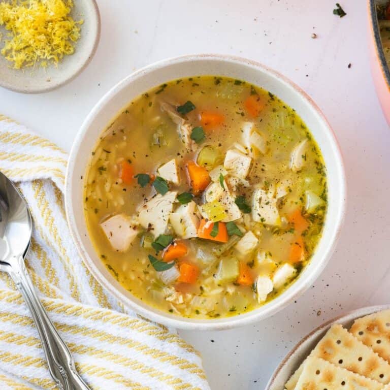 A bowl of lemon chicken orzo soup next to a yellow and white napkin with a spoon.