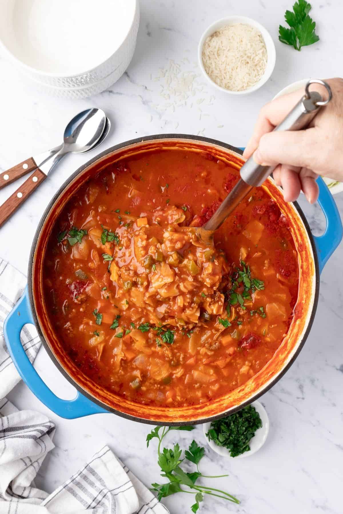 Unstuffed cabbage soup in a Dutch oven with a hand lifting up a ladle full.