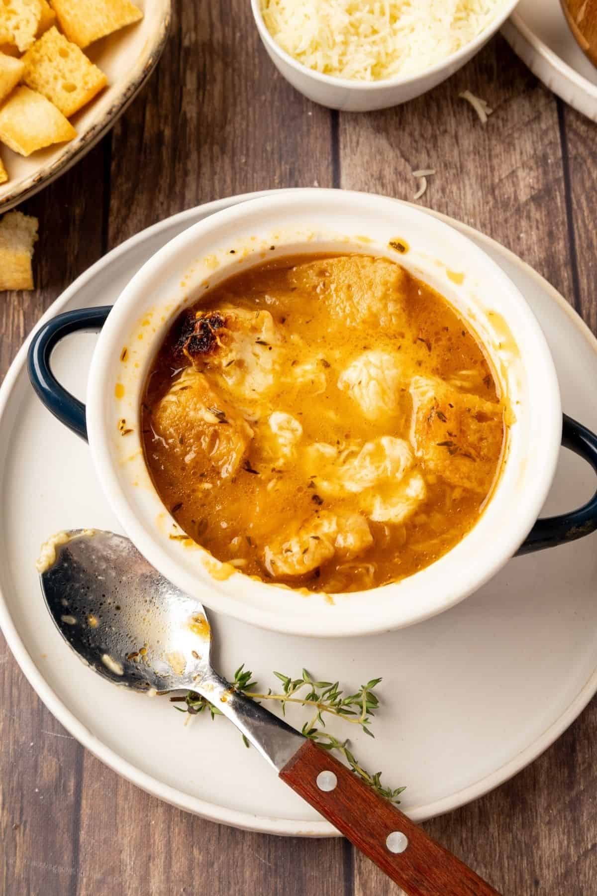 A bowl of Panera copycat French onion soup on a white plate with a spoon resting to the side.