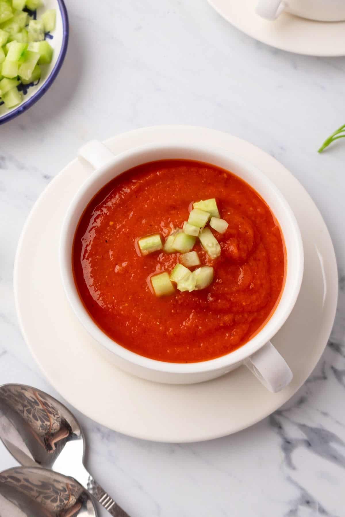 A bowl of cold tomato soup garnished with diced cucumber on a white plate. 