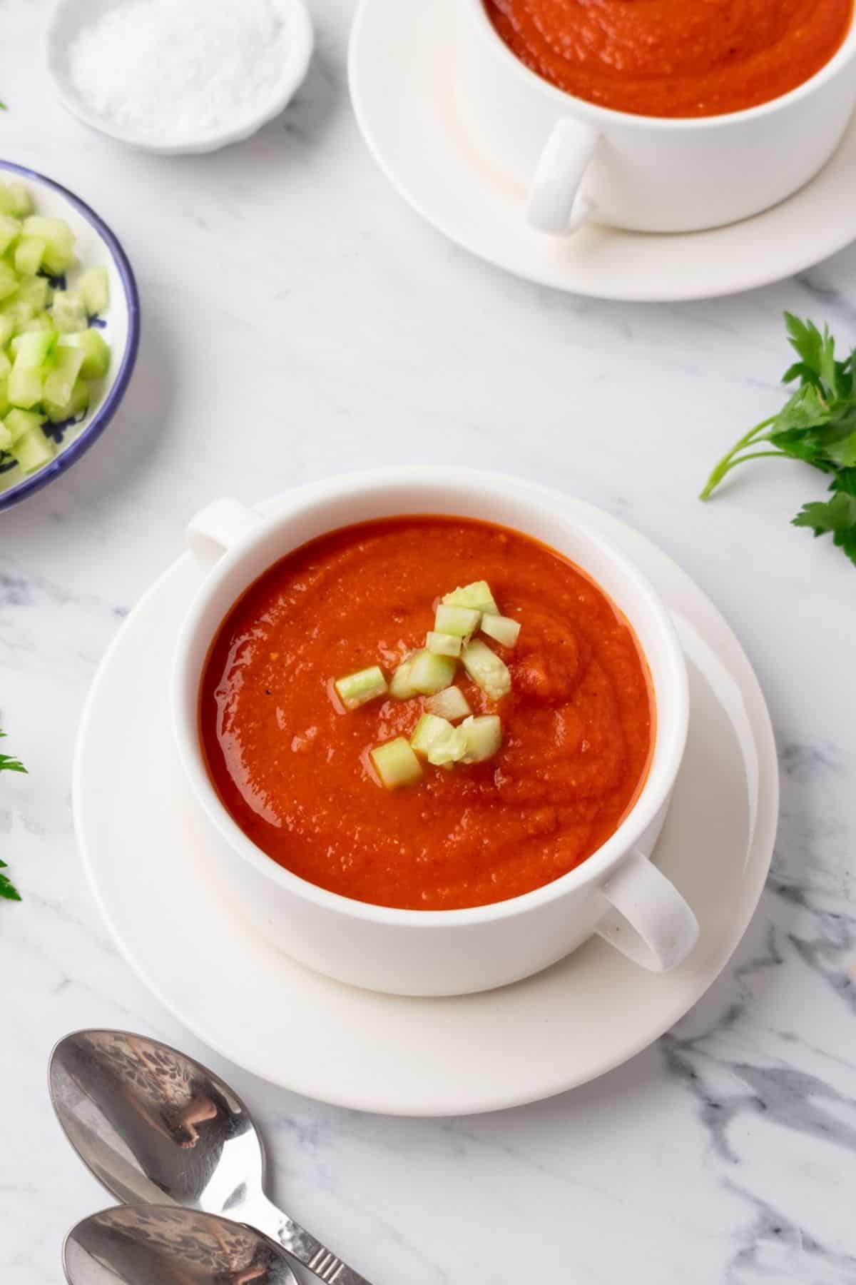 A bowl of chilled roasted tomato soup on the table topped with diced cucumber.