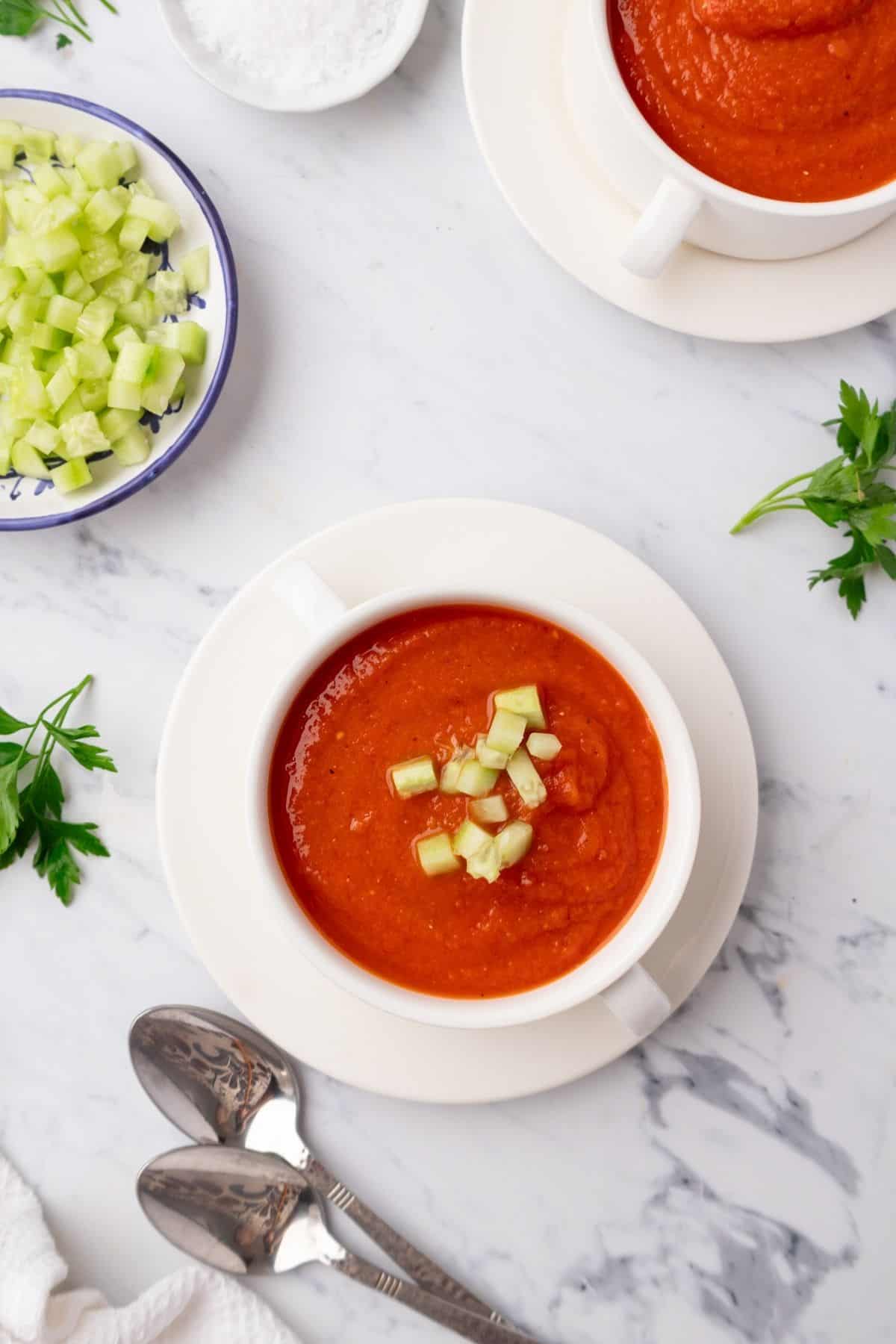 Bowls of cold tomato soup on a table with spoons and garnishes in bowls.