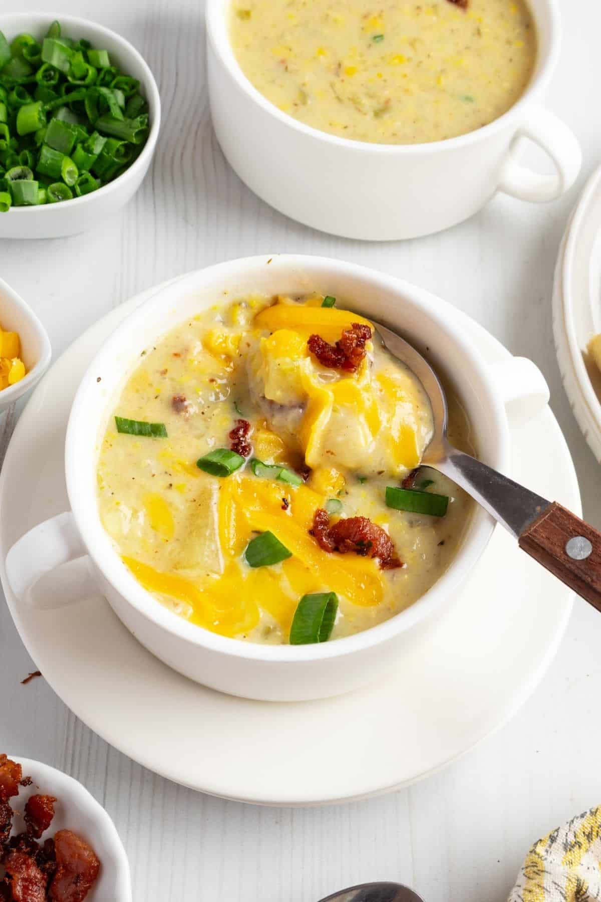 A bowl of potato corn chowder topped with cheese bacon and green onions and a spoon resting in the bowl.