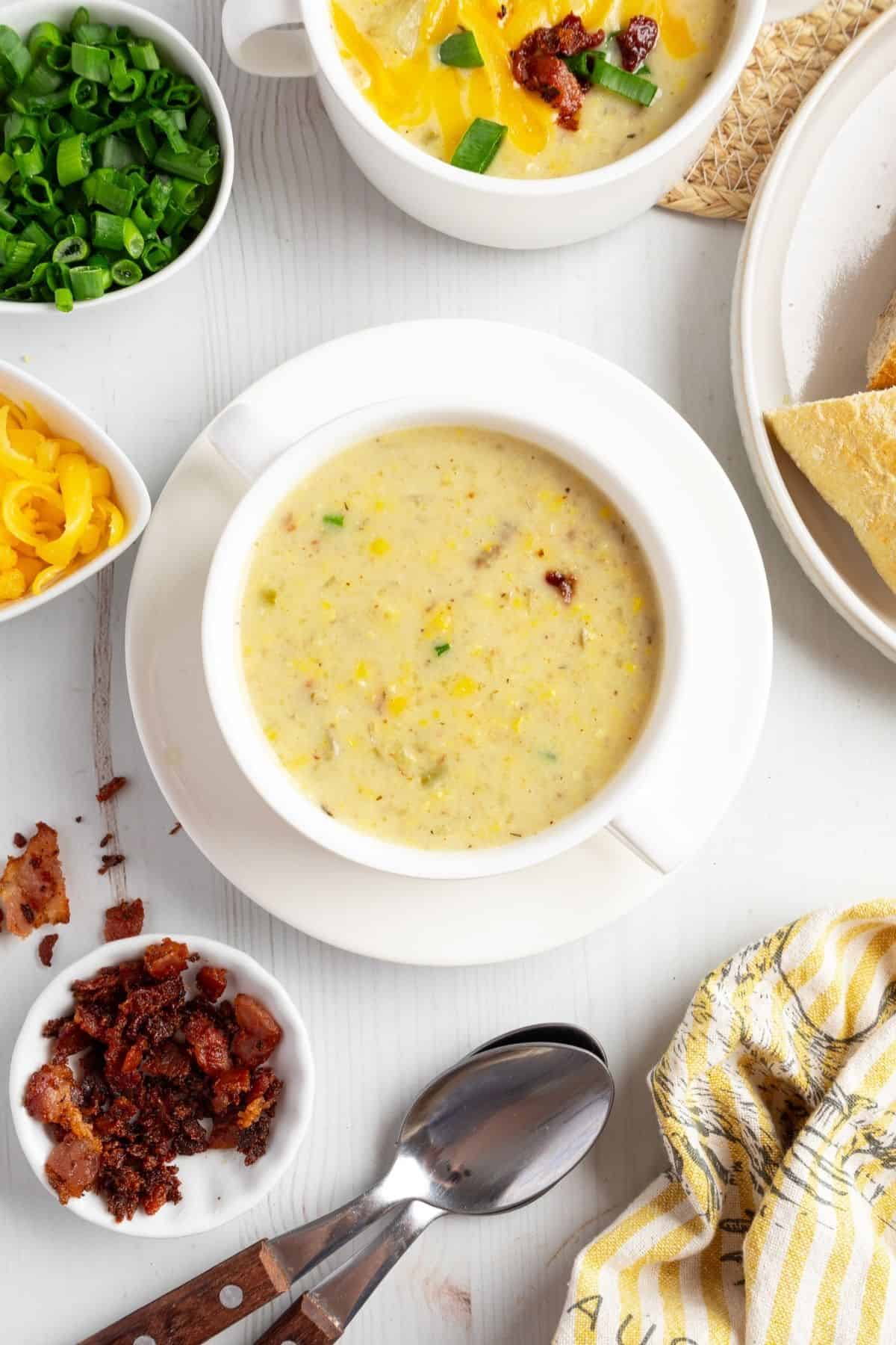 Creamy potato and corn chowder in a white bowl with bowls of bacon, green onions, and cheese around it.