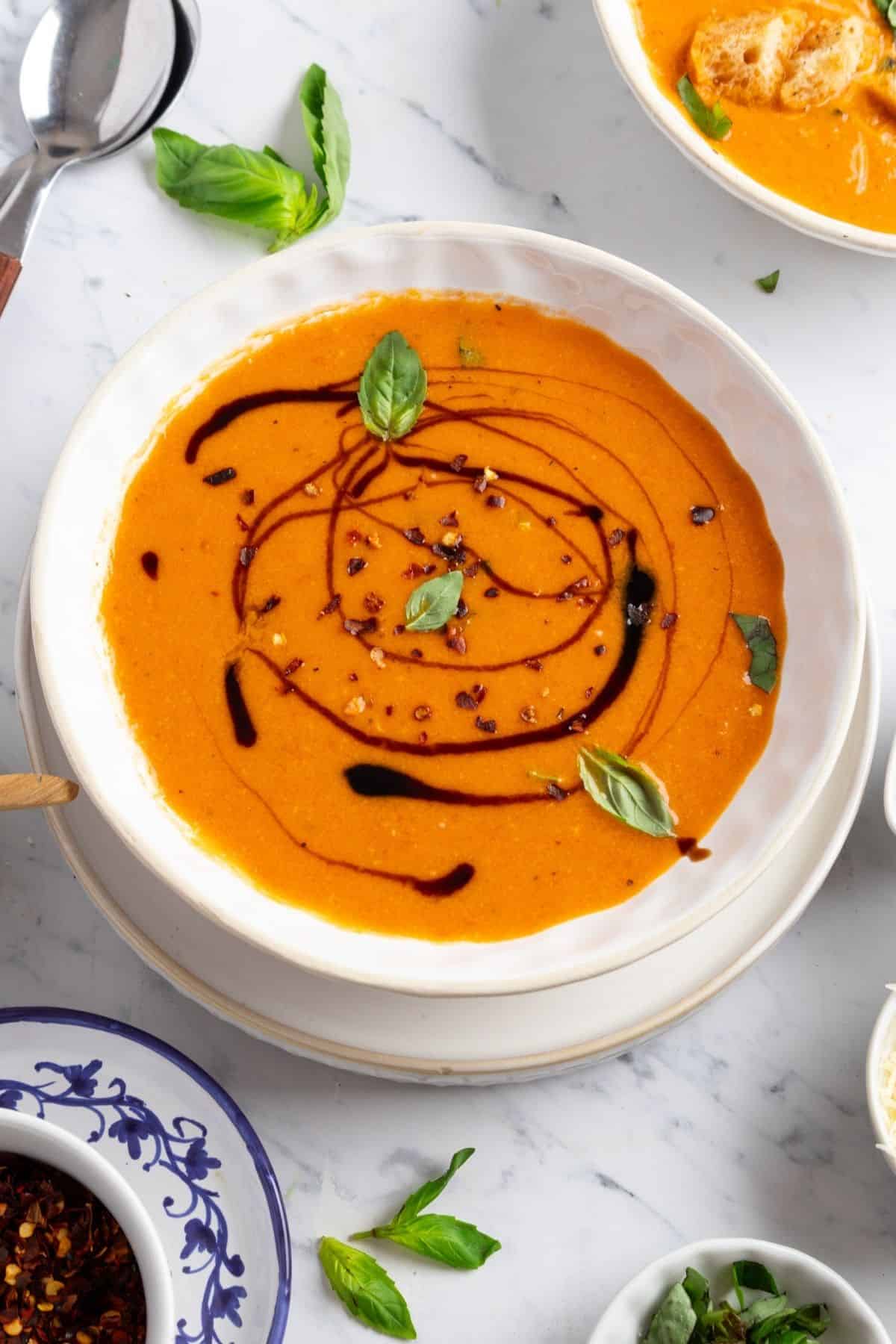 A bowl of roasted tomato basil soup drizzled with balsamic syrup and garnished with small basil leaves.