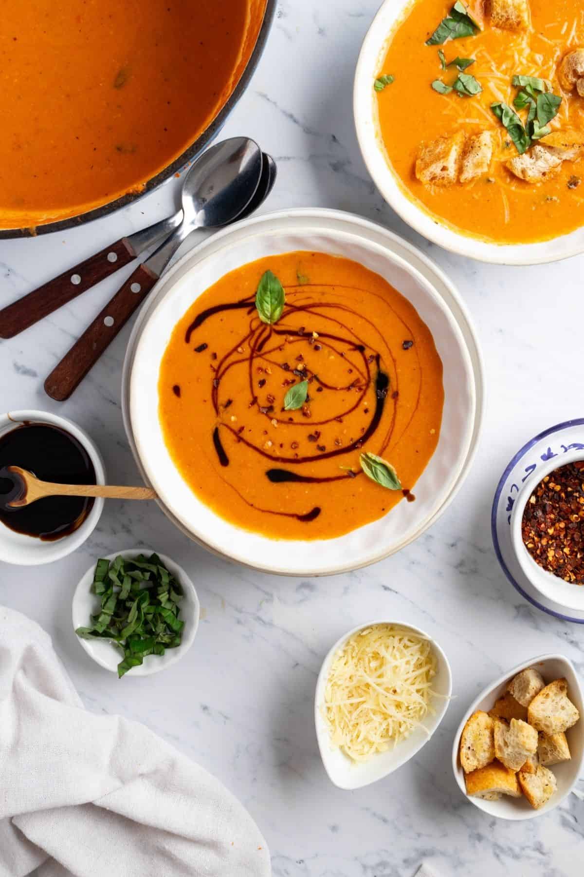 Bowls of roasted tomato basil soup on a table with bowls different toppings and the pot of more soup.