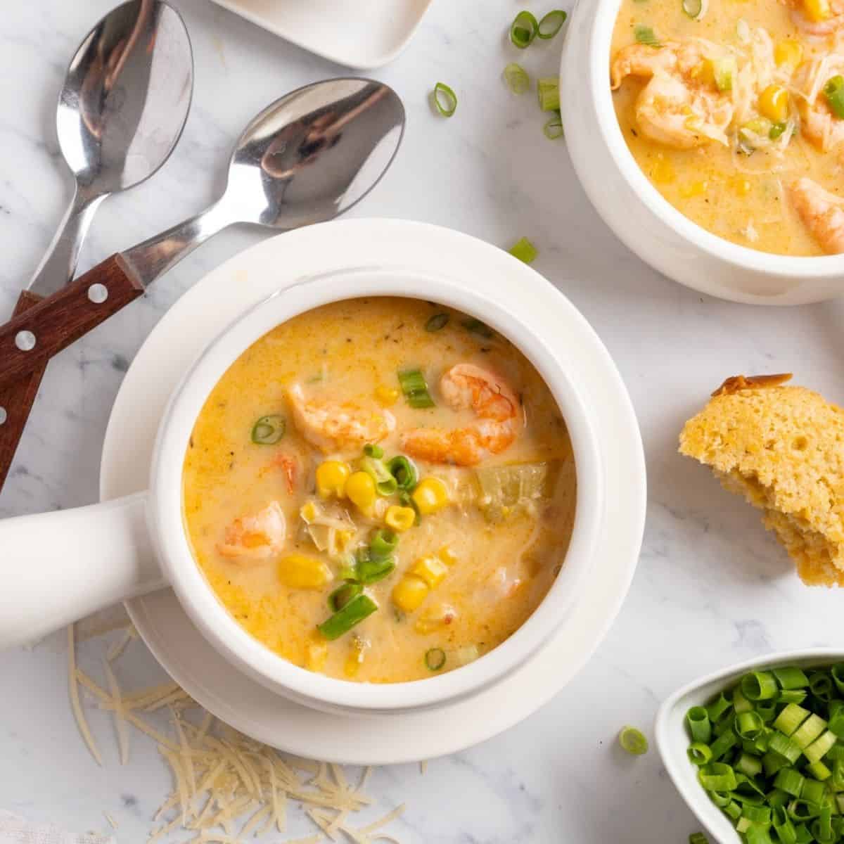 A white soup crock of shrimp corn chowder on a table with another bowl and toppings in the corners.